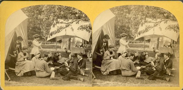 A group of people are gathered at a campsite next to a tent. A group of men and women are sitting on the ground playing cards, two women are standing, and another woman is sitting and sewing something she is holding in her lap.