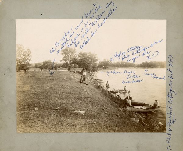 View along shoreline towards a group of men sitting at the edge of a lake, some of them in boats. The man on the far right sitting in a boat is Alex Baas. Karl Fauerbach wrote information on the photograph in blue ink, including, "Ed Brungelson milked his cows here + helped and so did his sister Nellies [sic]. They called her Cinderella."