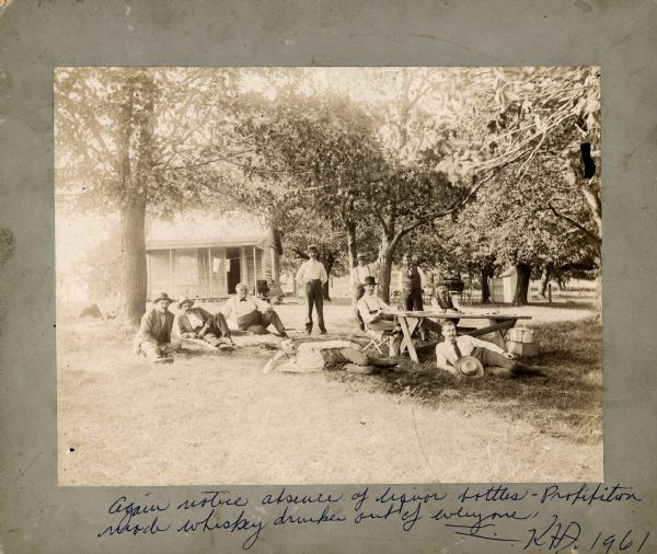 A group of friends relaxing outdoors. Some are lounging on the grass, two are sitting at a table, and some are standing. Alex Baas is sitting on the grass, third from the left. Philip Fauerbach is standing with his hands in his pockets, third from the right. On the bottom border of the image, Karl Fauerbach wrote the following in blue ink: "Again notice absence of liquor bottle. Prohibition made whiskey drunks out of everyone."