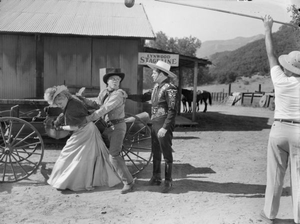 Outdoor Set of the TV show "Cisco Kid." The Cisco Kid has his hand on the shoulder of a man who is is holding onto a woman. A man holding a microphone boom is on the right.
