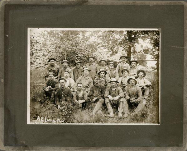Outdoor group portrait of the miners who work at Optimal Mine. They are seated outdoors on the grass, and are wearing work clothes and hats.