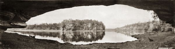 Panoramic view of the Wisconsin River in the Wisconsin Dells from underneath an overhanging rocky outcropping. A canoe is beached on the far left. The words "UNDER THE OVERHANGING ROCK, WISCONSIN DELLS. Copyright 1900. by H.H. Bennett." are on a label on the right.