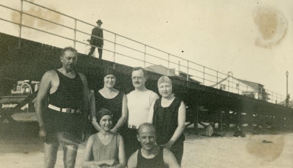 Group portrait of Alvin Kraenzlein with two other men and three women. They are all wearing bathing suits and posing on a beach. The women are wearing bathing caps. Behind and above them is a boardwalk.