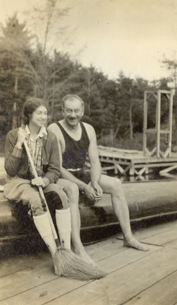 Portrait of Alvin Kraenzlein and his daughter, Claudine, sitting outdoors together on an overturned boat on a dock. She is holding a broom.