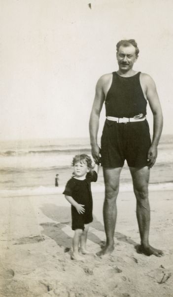 Alvin Kraenzlein standing on a sandy beach holding the hand of his toddler son. Behind them is the shoreline.
