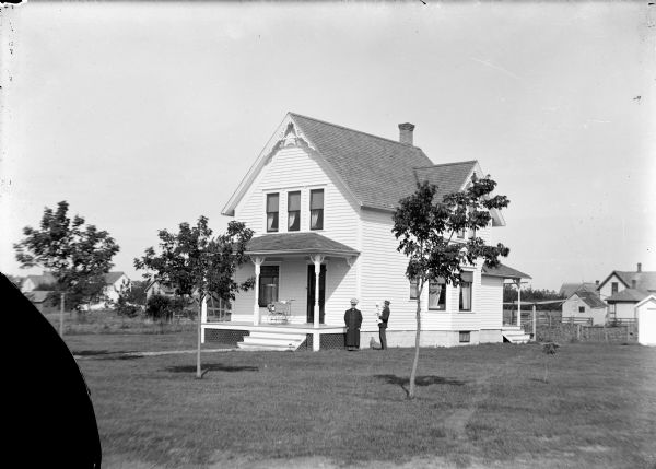 View across lawn towards two people standing outdoors in front of a house. Three trees are planted in the front yard. Other buildings are in the background on the left and right.