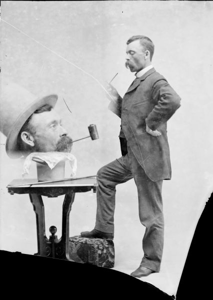 Trick photographic portrait of a man standing next to a table and holding a pipe in his hand. On the table is a man's large head placed on a pedestal. He is also smoking a pipe. 
