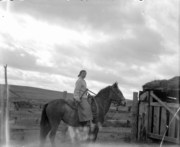 Woman on horseback posing near a fence. There is a hill in the background.