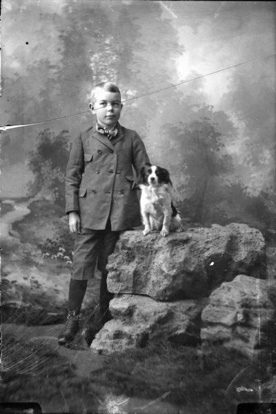 Full-length studio portrait in front of a painted backdrop of a boy posing with a small dog. The dog is seated on a prop rock formation. The boy is wearing a suit coat, short pants, and knee socks.