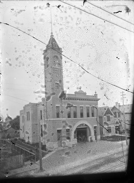 Elevated view from across the street of the new North Side Fire Station, Fire Station No. 1, at 412 N. 8th Street.  The construction is two-story brick, has a decorative stone arch around the garage door, corbeled cornice and a hose tower on the left corner.  A sign above the door reads:"Fire Station No. 1."  Houses are on the far right.  A newspaper article regarding plans for the new station dated August 13,1901 in the Manitowoc Daily Herald states:  "The structure is to be built on 8th Street near Chicago on property purchased from J. Kucera some years ago.  It will be of solid brick, with Bedford stone trimming, stone foundation, two stories high with basement and its dimensions are 98 by 32 feet.  At the southeast corner there will be a hose tower 80 feet high, which with the front of the building will be of pressed brick.  The first floor will be taken up by an apparatus room 80x65 feet large, with stables in the rear, containing six regular stalls and a box stall.  On the second floor will be the chief's office, men's sitting room, bedrooms, bathrooms and hayloft.  The first story will be finished in white and the second story in yellow pine.  Architect W. J. Raeuber of this city drew the plans."  The Sanborn Map Company's 1906 insurance map indicates the fire station is two-story brick with twelve-inch-thick walls, thirty feet in height with a fifty-foot tower.  It has ten men, five horses, one hose wagon, 2000 feet of 2 1/2 inch hose and four Babcocks.  The city's South Side Fire Station, Fire Station No. 2, is at 911 Franklin Street.