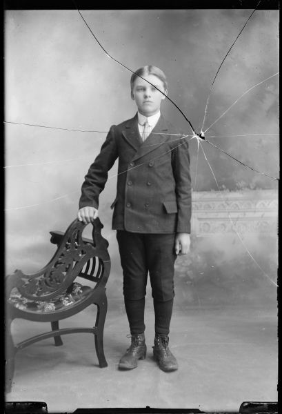 Full-length studio portrait in front of a painted background of Vesey Vernon. He is standing with his hand resting on a chair and is wearing a suit coat, necktie, and short pants.