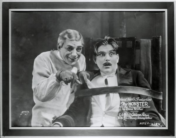 Dr. Ziska (Lon Chaney) standing next to Amos Rugg (Hallam Cooley) who is strapped into a chair in a scene for the film "The Monster." Rugg is dressed in a tuxedo and has a surprised look on his face; Dr. Ziska is smiling.