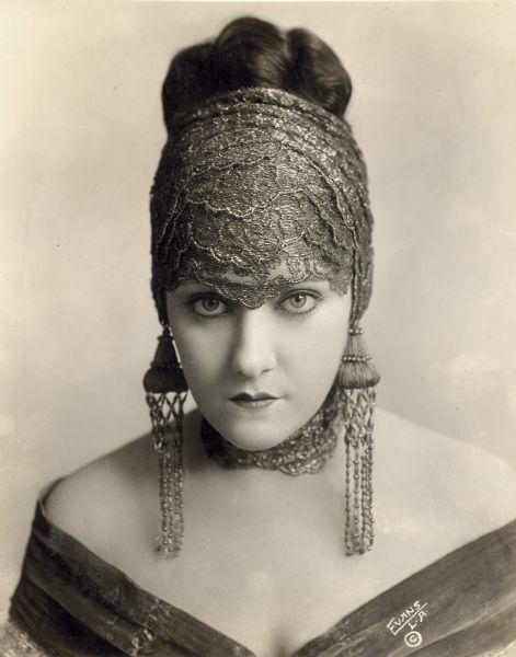 Publicity portrait of Gloria Swanson. She is looking straight at the camera and is wearing an off the shoulder dress, large dangling earrings, and a headpiece or scarf that covers her forehead, ears, and neck.