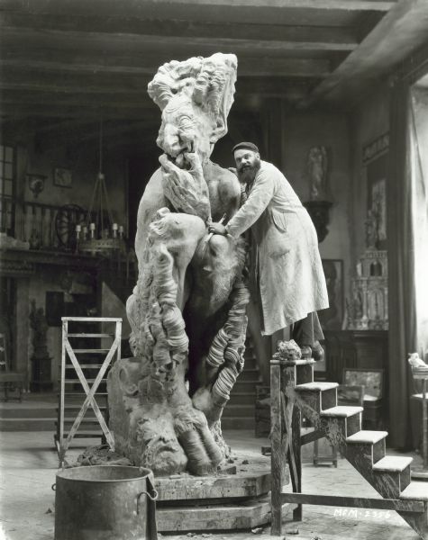 An artist posing with a large statue that was used in the film "Ben-Hur: A Tale of Christ."  The artist is standing on the top step of a wooden staircase and is resting his hands on the statue.