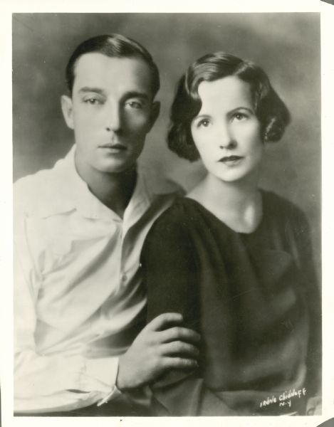 Waist-up portrait of Buster Keaton and his wife Natalie Talmadge sitting together. He is sitting on the left and is holding her arm.