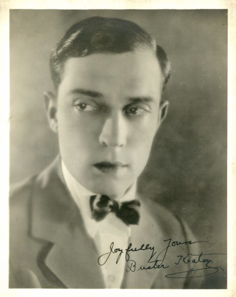 Quarter-length portrait of a young Buster Keaton. He is wearing a suit and bow tie and is looking off to the left. Autographed on the bottom right: "Joyfully Yours Buster Keaton."