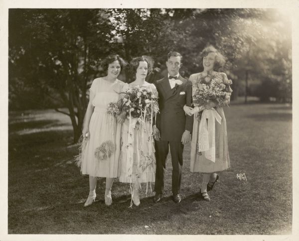 Norma, Natalie, and Constance Talmadge standing outdoors with Buster Keaton on the day he married Natalie. Natalie and Constance are both holding big bouquets of flowers. Buster has a blank expression on his face.