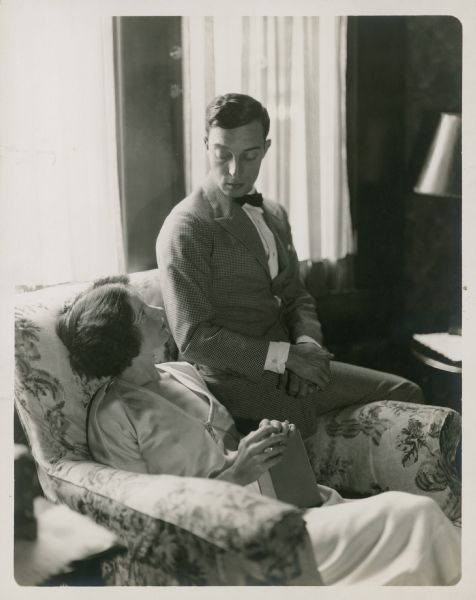 Buster Keaton sitting on the arm of an oversize chair looking down at his wife, Natalie, who is sitting in the chair looking up at him. She is holding a book. The photograph is from their honeymoon.
