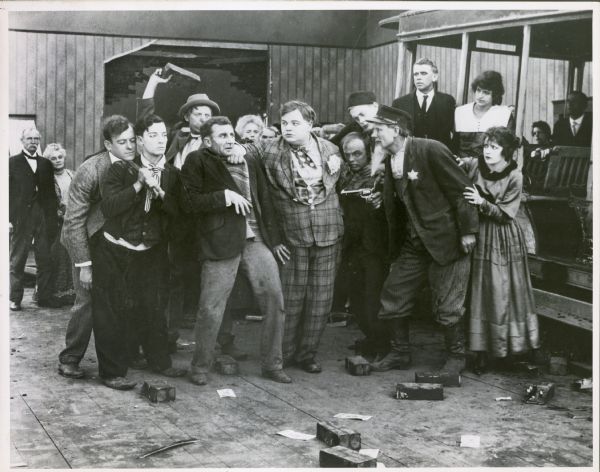 Scene still from the 1918 silent film <i>The Bell Boy</i>. Roscoe "Fatty" Arbuckle is standing at the center of a group, wearing a plaid suit. He is holding onto a man that other people want to attack. Buster Keaton is standing on the far left next to the man, while holding onto the tie of the man behind him.
