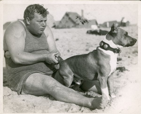 Roscoe "Fatty" Arbuckle sitting on a beach wearing a swimsuit and holding the tail of Luke the dog.