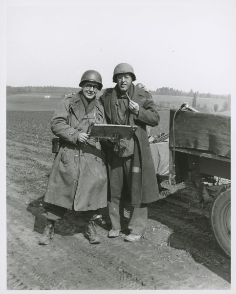 John MacVane (left) and another man standing together in a field next to a military vehicle. They are both wearing military garb, including long coats and protective helmets. The man on the left is holding something in his hands, and appears to have a set of binoculars in a case on his waist.