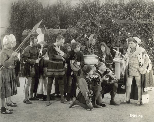 William Farnum, as Francois Villon, speaking to a crowd of people in a still for the film "If I Were King." Two men are kneeling before him, while the rest of the men are standing. There are four men dressed as soldiers holding large spears.
