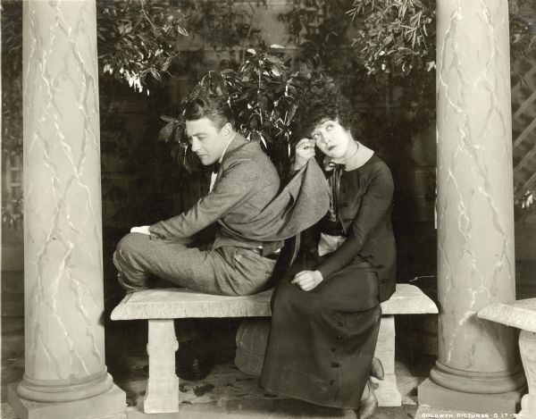 Tom Moore and Mabel Normand are sitting next to each other on a bench in a still from the film "The Floor Below." Normand is drying her eye with Moore's coattail as he is looking away. They are sitting between two stone pillars.