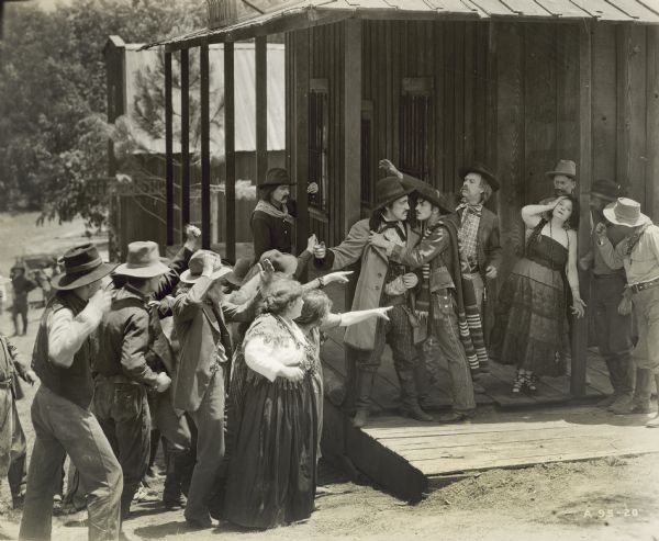 Still from the film "Scarlet Days." A group of men and women are standing on a street and pointing toward a woman who is leaning against a pole. A number of men are standingc
 near her. The woman has one hand on her head and is wearing a strapless dress.