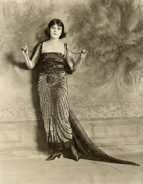 Theda Bara posing in front of a wall or backdrop that looks like a forest. She is holding strands of beads, which are attached to her dress, in each hand. The dress also has a sheer train.  