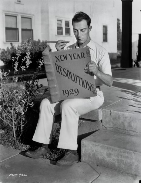 Buster Keaton sitting outdoors and holding an oversize book and pencil. The book is for New Year Resolutions in 1929.