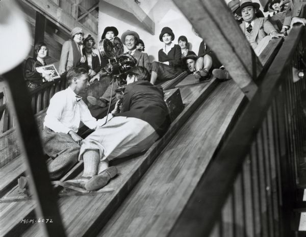 Production still from the 1928 film "The Crowd." Two cameramen are sitting on a wooden slide behind a small camera. People are sitting at the top of the slide waiting to go down.