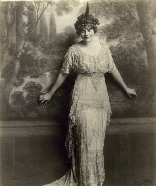 Publicity photo of actress Myrtle Stedman. She is wearing a lace dress and a headpiece with one feather. She is standing in front of a wall made to look like a forest and is touching the wall behind her with her hands.