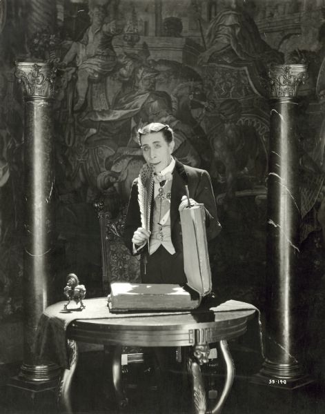George Arliss, as Dr. Muller, standing behind a table with a large book in a still for the film "The Devil." He is holding a large quill pen in one hand up to his mouth. There is a column on each side of the table.