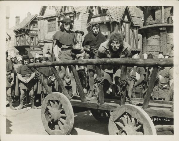 The hunchback (Lon Chaney) standing in a cart with two men standing behind him in a still for the film "The Hunchback of Notre Dame." One man is holding an hourglass.  