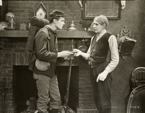 Thaddeus Briggs (Spottiswoode Aitken) is handing his grandson Ulysses S. Grant Briggs (Charles Ray) something in a still for the 1919 film "Hay Foot, Straw Foot." Ulysses is dressed as a soldier and is holding a rifle in his other hand.