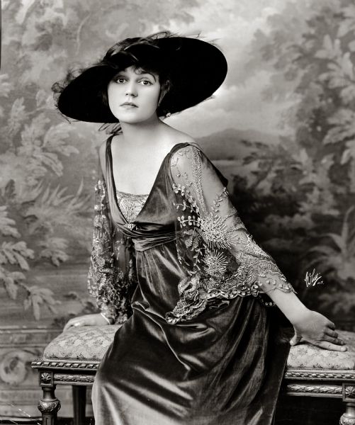 Alice Brady posing for a publicity photograph while sitting on a cushioned seat and leaning in one direction. She is wearing a wide brimmed hat and a dress with sheer embroidered sleeves.