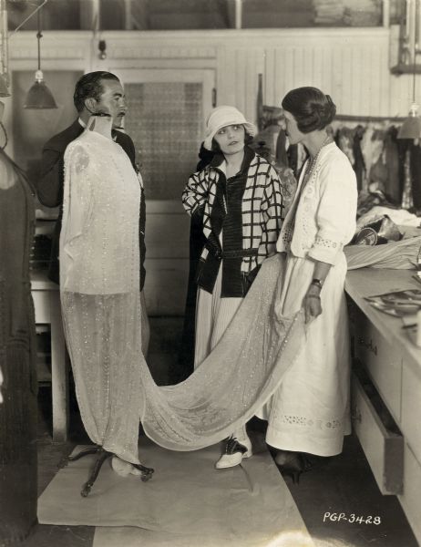 Producer George Fitzmaurice and actress Pola Negri talking with Ethel Chaffin, chief costume designer for Lasky Studio, about Negri's costumes for the 1923 film "Bella Donna." Chaffin is holding the train of a dress that is on a wardrobe form.