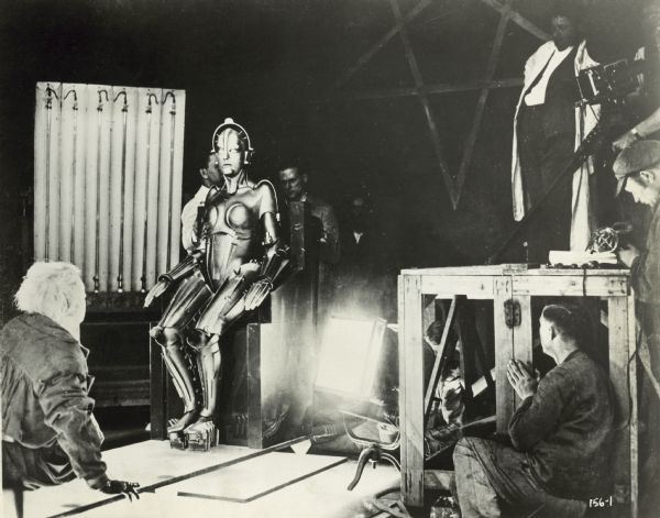 A behind the scenes photograph from the set of the film "Metropolis. "Men are seen behind and below where the Maria robot is sitting. The camera can be seen at the top right of the image.
