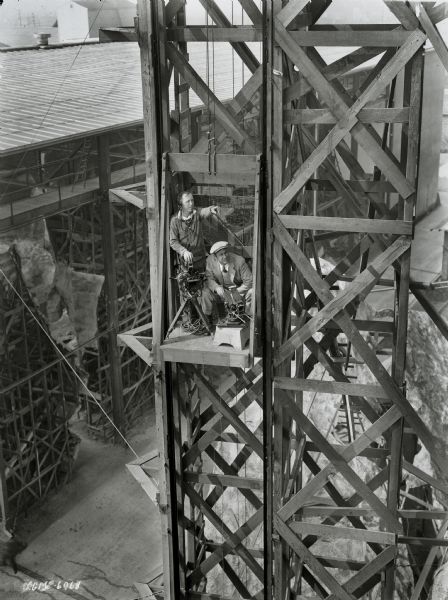 Cinematographers Reggie Lanning and Elgin Lessley sitting and standing on a small platform with two cameras during the filming of Buster Keaton's movie "The Cameraman." The platform is part of a wooden scaffolding and can be moved up and down. Lanning is pointing towards something.