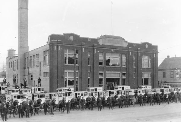 Elevated view of horse-drawn milk wagons and drivers posing in front of the Kennedy Dairy Company, 621-629 West Washington Avenue. Some drivers are standing on the roofs of their wagons. The Kennedy Dairy Company was a two-story brick building with large windows. Two men are looking out of the large second-story window above the entrance.