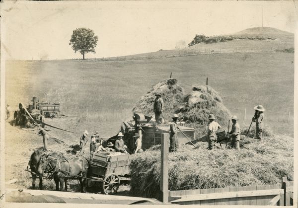 Elevated view of men harvesting what appears to be hay on the Lloyd Jones farm. A tractor in the background on the left is powering the thresher with a long belt. Men are standing on and around a horse-drawn wagon which is backed up to the thresher.
