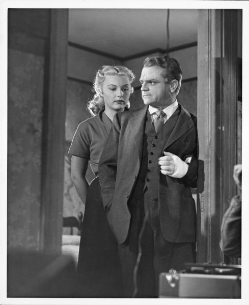 Publicity still of Barbara Payton and James Cagney from the film "Kiss Tomorrow Goodbye." Payton is standing just behind Cagney and is looking over his shoulder. Cagney's hand is wrapped in a bandage.
