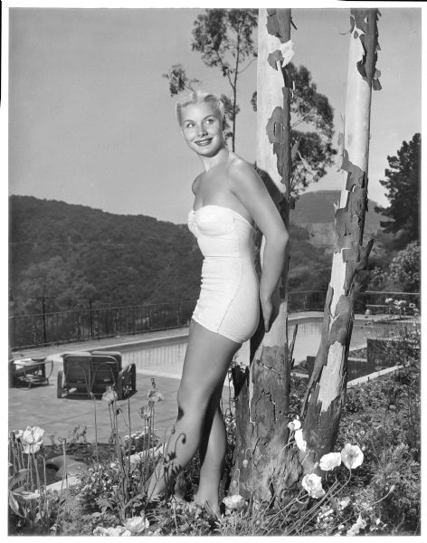Barbara Payton posing outdoors for a publicity portrait for the film "Trapped." She is standing against a tree wearing a swimsuit. A swimming pool is below in the background.
