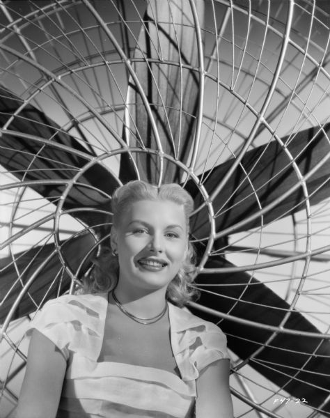 Head and shoulder portrait of Barbara Payton sitting in front of a large fan in a publicity portrait for the film "Trapped." She is wearing a white dress and a double stranded necklace.
