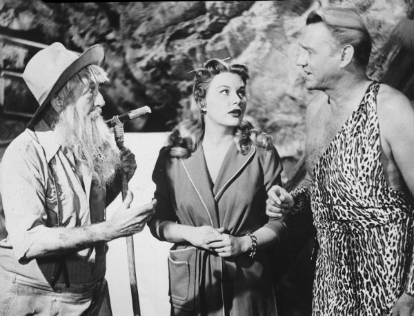 Publicity still of Barbara Payton, Sonny Tufts, and another man in the film "Run for the Hills." Tufts is wearing a leopard print tunic, and Payton is wearing a house coat. The other man has a long scraggly beard, and is wearing a hat and holding a walking stick.