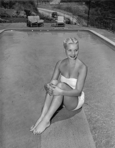 Publicity still for the film "Trapped." Barbara Payton is wearing a white bathing suit and sitting on a diving board above a swimming pool.