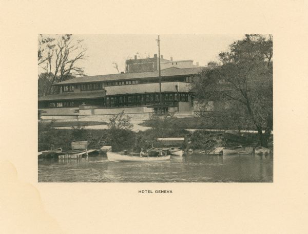 A view across water towards the Lake Geneva Hotel, also called Hotel Geneva. There are several boats and piers at the water's edge. Two men are boating in the foreground.