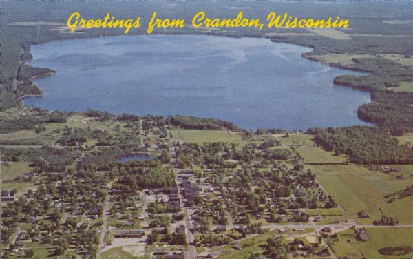 Aerial view of town, showing all of Lake Metonga in the background. Caption reads: "Greetings from Crandon, Wisconsin."