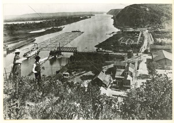 A woman and a man are standing on a hill overlooking a large lumber raft (allegedly the last of its kind) traveling down the Mississippi River. There is a tow boat behind the raft and a bow boat in front of it, which is used for steering the raft. The boats and raft are passing a pontoon bridge, which is opposite Prairie du Chien, Wisconsin.