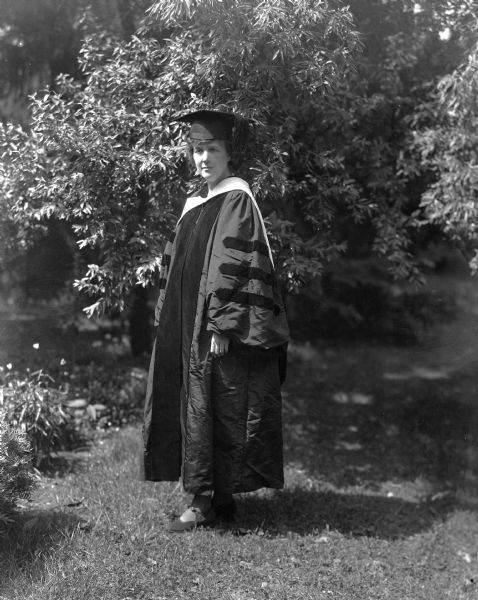 Zona Gale Breese (1874-1938) posing outdoors for a full-length portrait in doctoral academic cap and gown on the occasion of receiving an honorary Doctor of Literature degree from the University of Wisconsin.